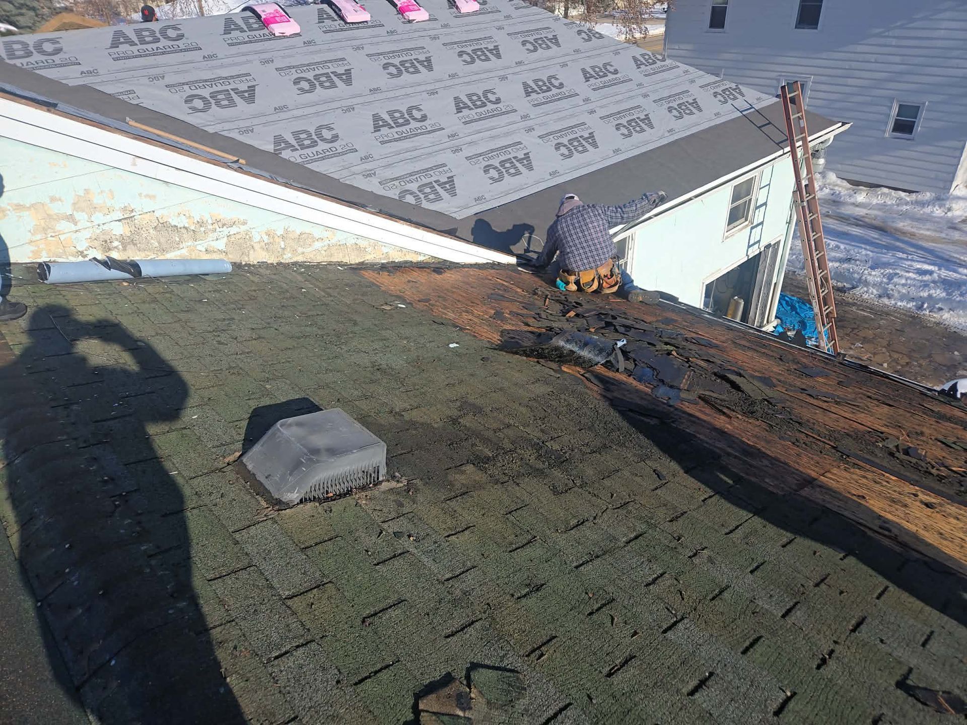 Roof being repaired, showing old and new shingles. Worker on the roof, partially covered in shadow.