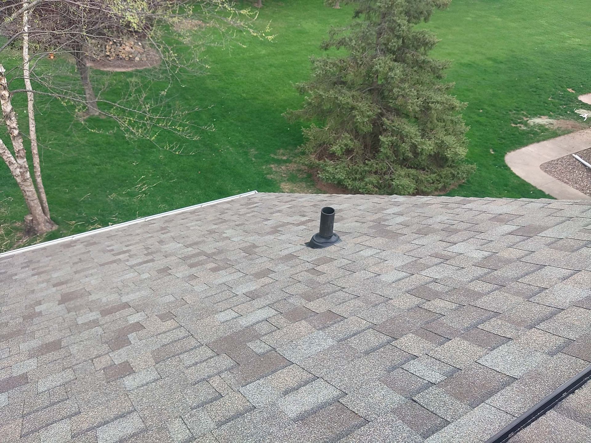 A roof with gray shingles and a black vent pipe, overlooking a grassy yard with trees.