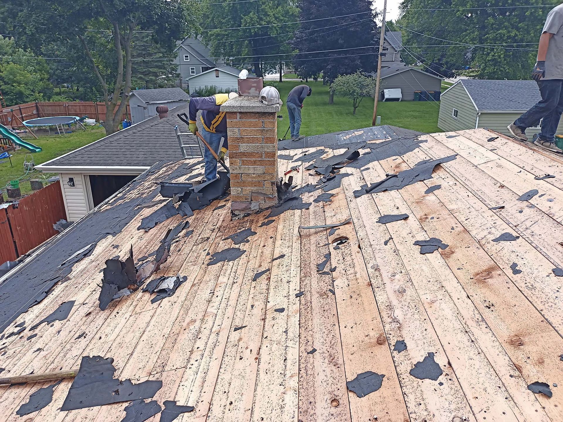 Workers removing old shingles from a roof, focusing around a brick chimney.