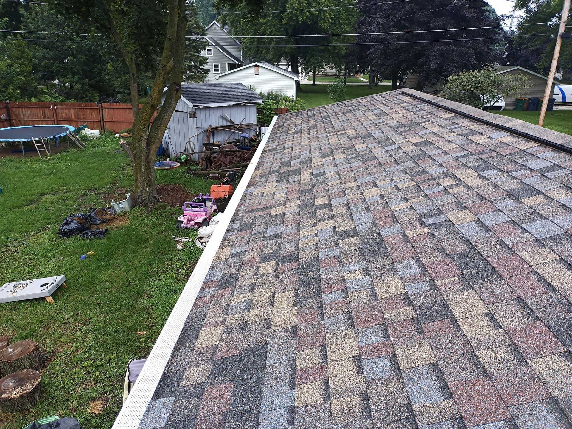 View of a roof with brown and grey shingles, white gutter, and backyard with grass, trees, and shed.