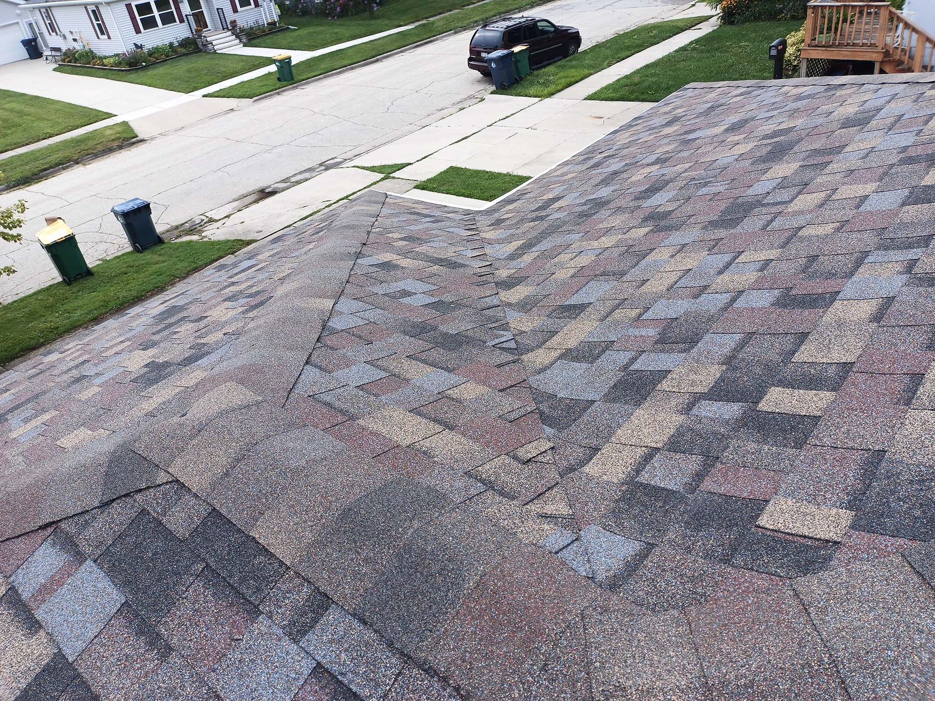 A close-up view of an asphalt shingle roof. The roof is weathered with a mix of colors. In the background, a street and houses are visible.