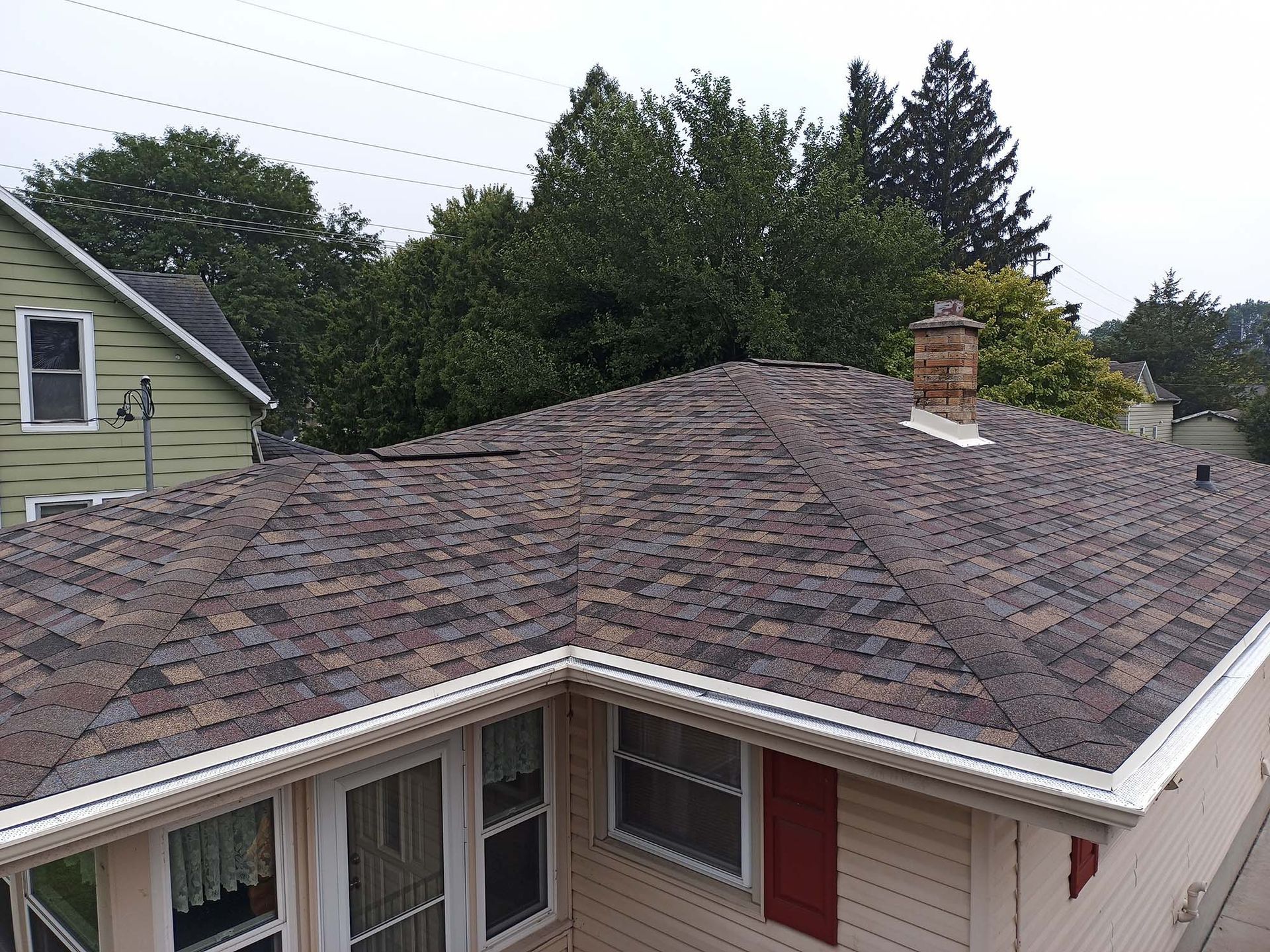 A house roof with brown shingles, white gutters, and a brick chimney, with trees in the background.