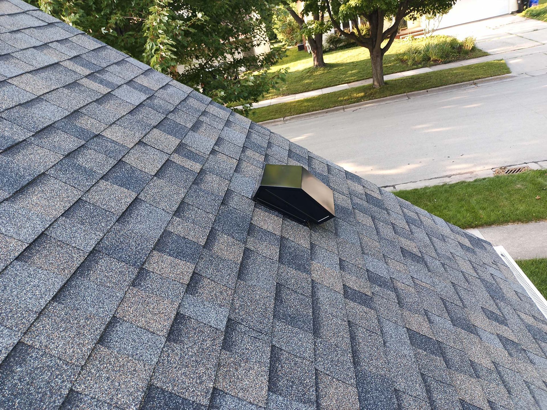 Dark gray asphalt shingle roof with black vent, overlooking street and trees.