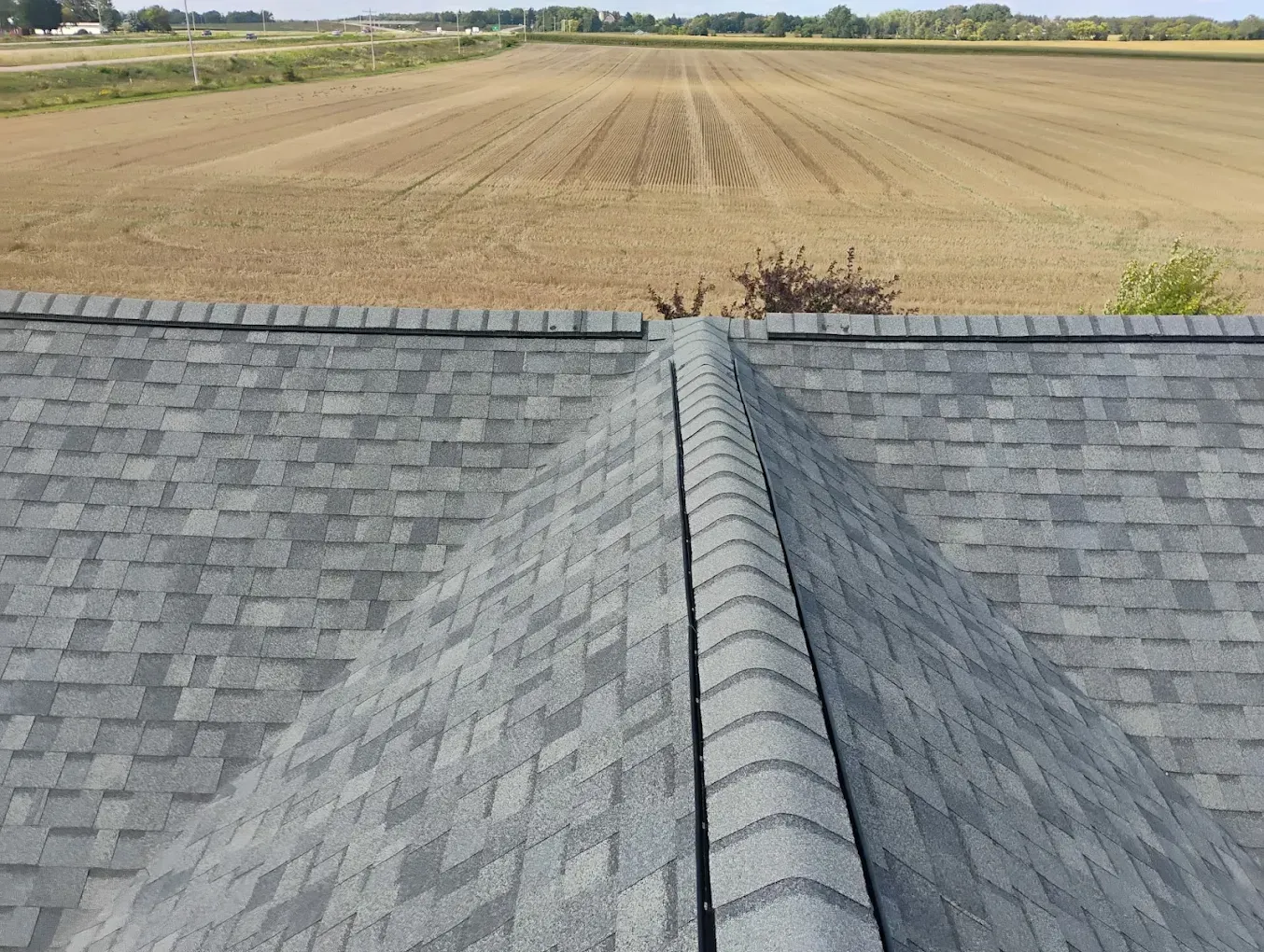 Gray shingle roof peak with a field in the background under a sunny sky.