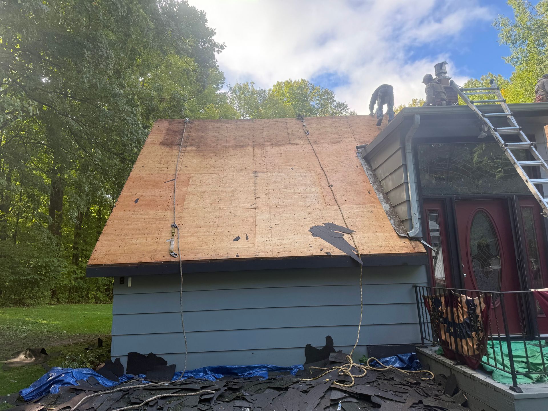 Roofers on a house roof partially covered with brown underlayment. A ladder, green grass, and trees are visible.