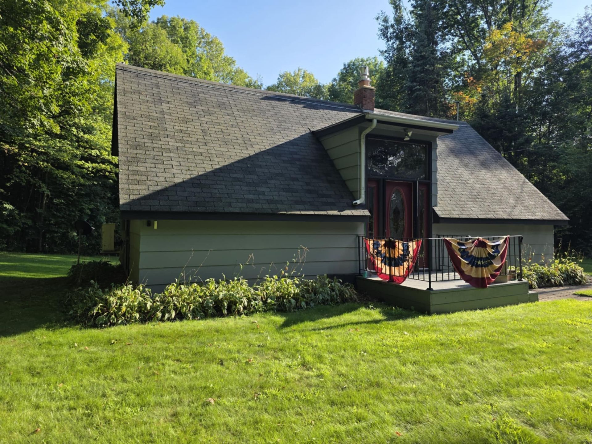 Cottage-style house with green roof and walls, small porch with decorative flags, surrounded by green grass and trees.