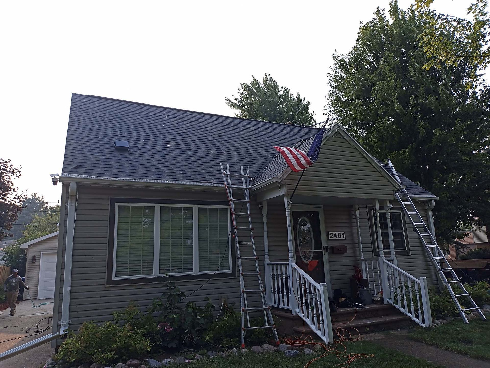 House with new dark shingle roof, ladders, American flag, and tan siding.