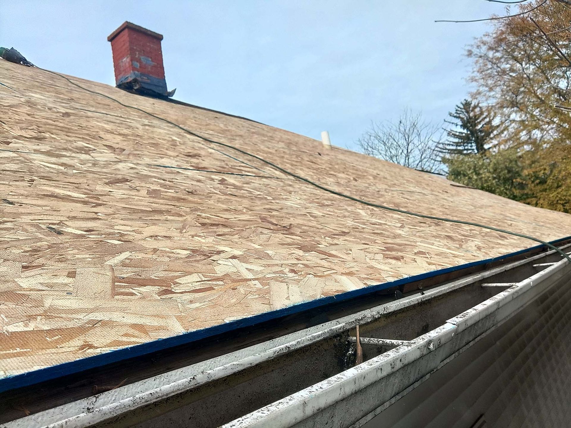 Roof with exposed wooden sheathing, gutters, and a chimney against a blue sky.