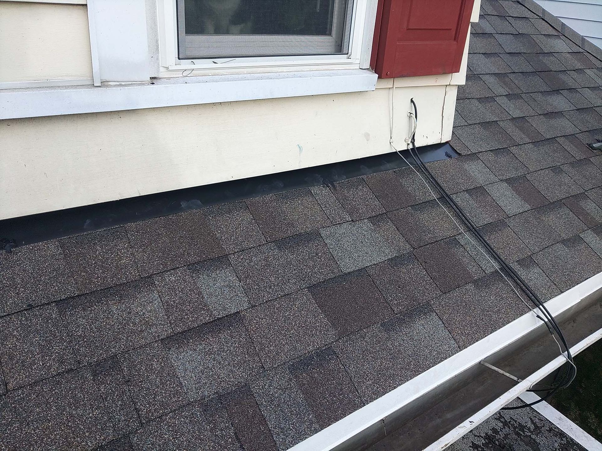 Brown shingle roof next to a beige house wall with a window and a gutter.