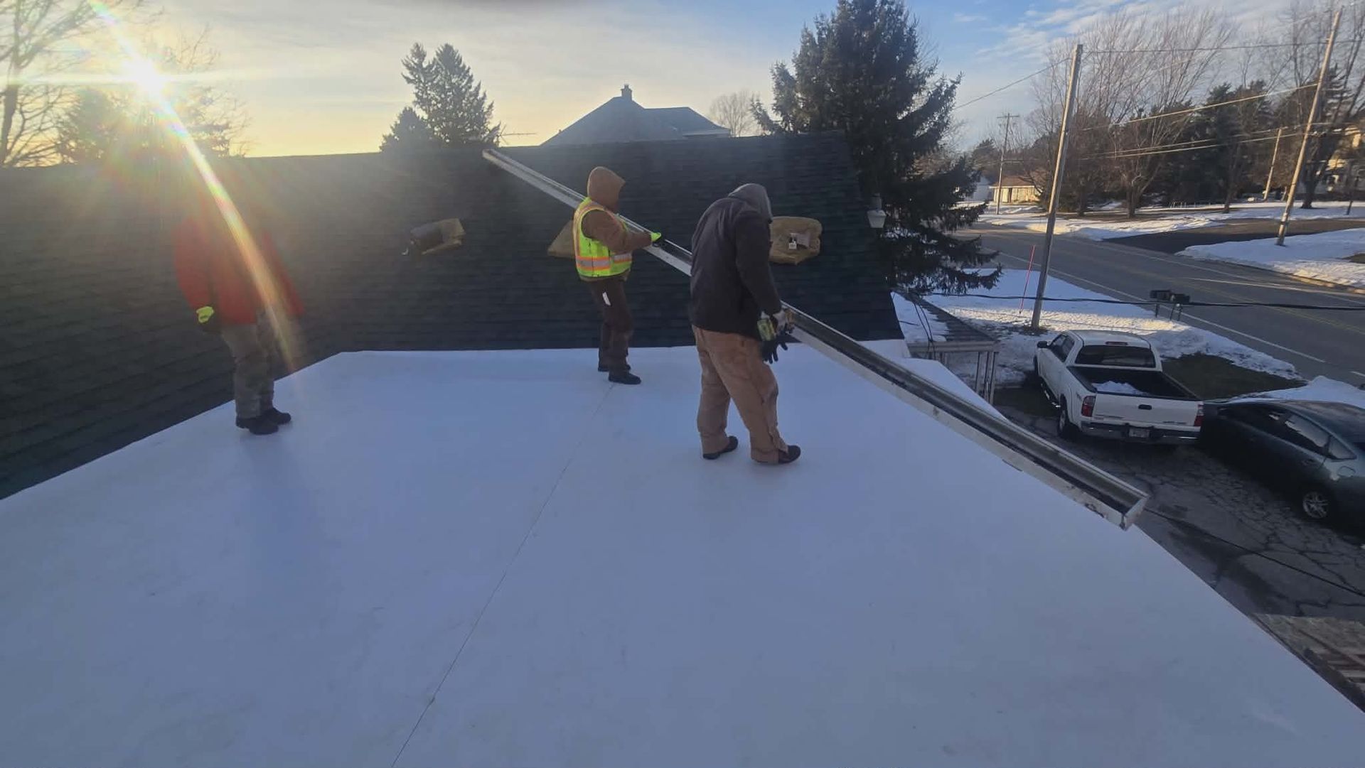 Three workers installing roofing material on a rooftop, with a sunny backdrop and parked vehicles nearby.