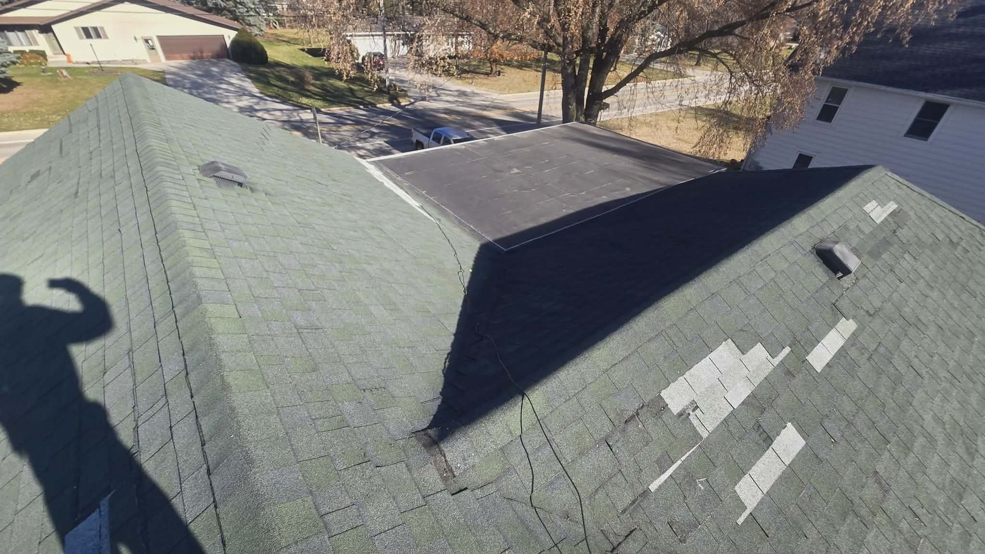 View of damaged asphalt shingle roof. Patches missing. Nearby are other buildings and a street.