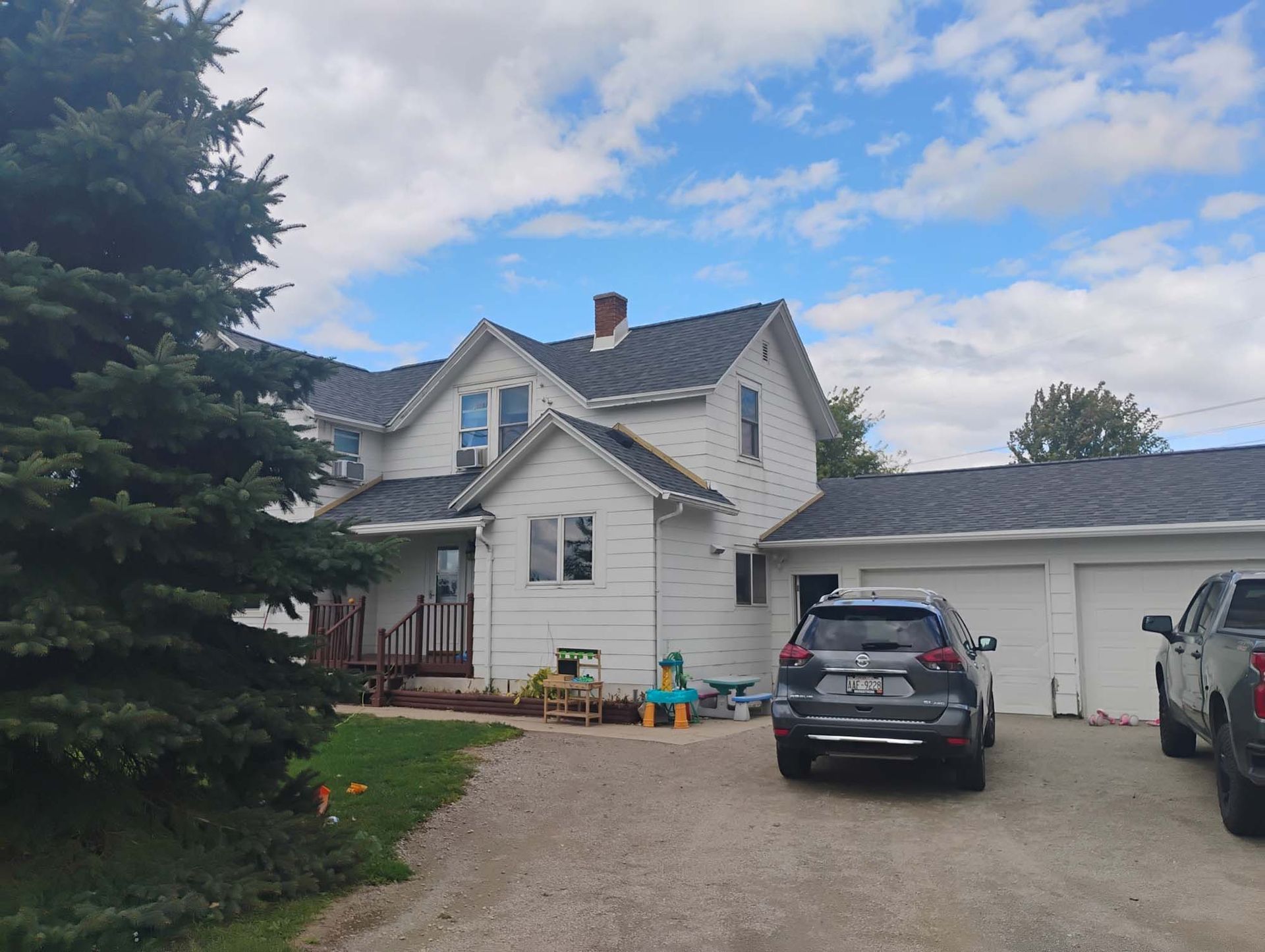 White two-story house with detached garage; gravel driveway; blue sky with clouds; parked cars.