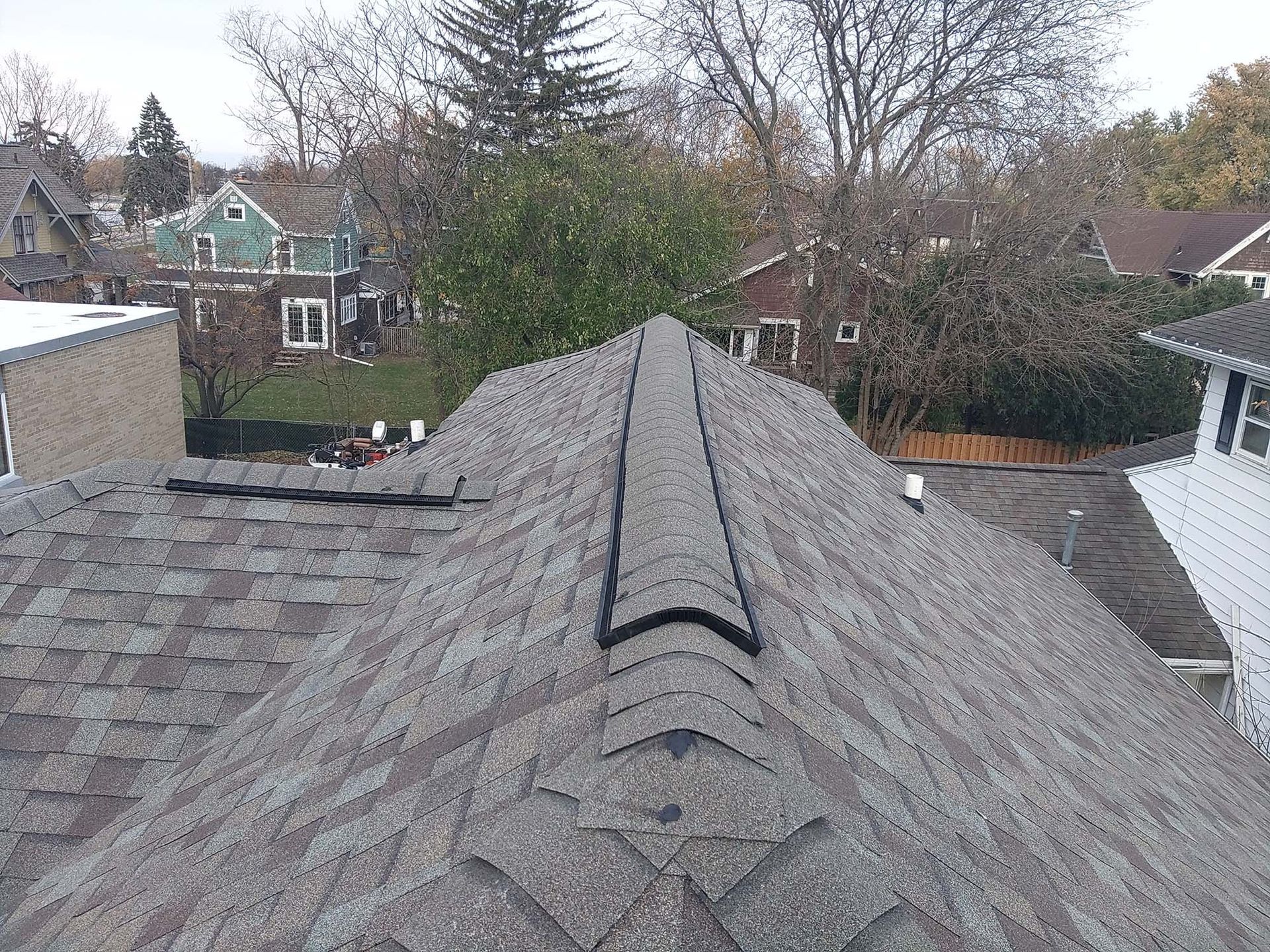Rooftop with gray shingles and a dark ridge cap, with houses and trees in the background.