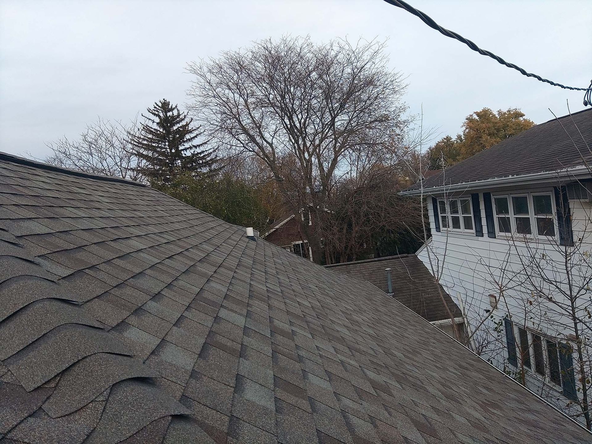 View of a brown shingled roof, trees, and the side of a white house with windows under a cloudy sky.
