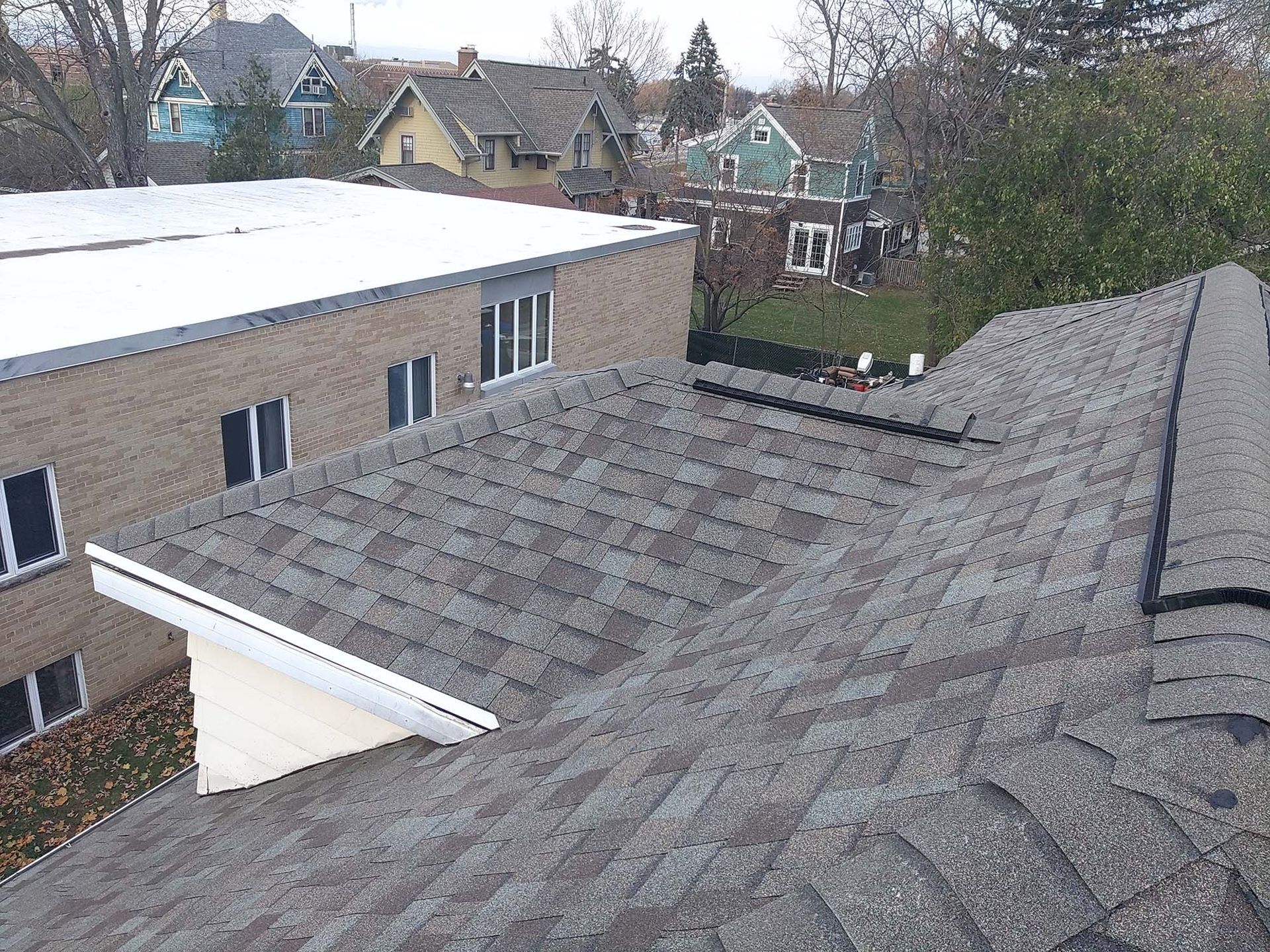 View of a gray shingled roof with a flat roof building behind it, residential houses in background.