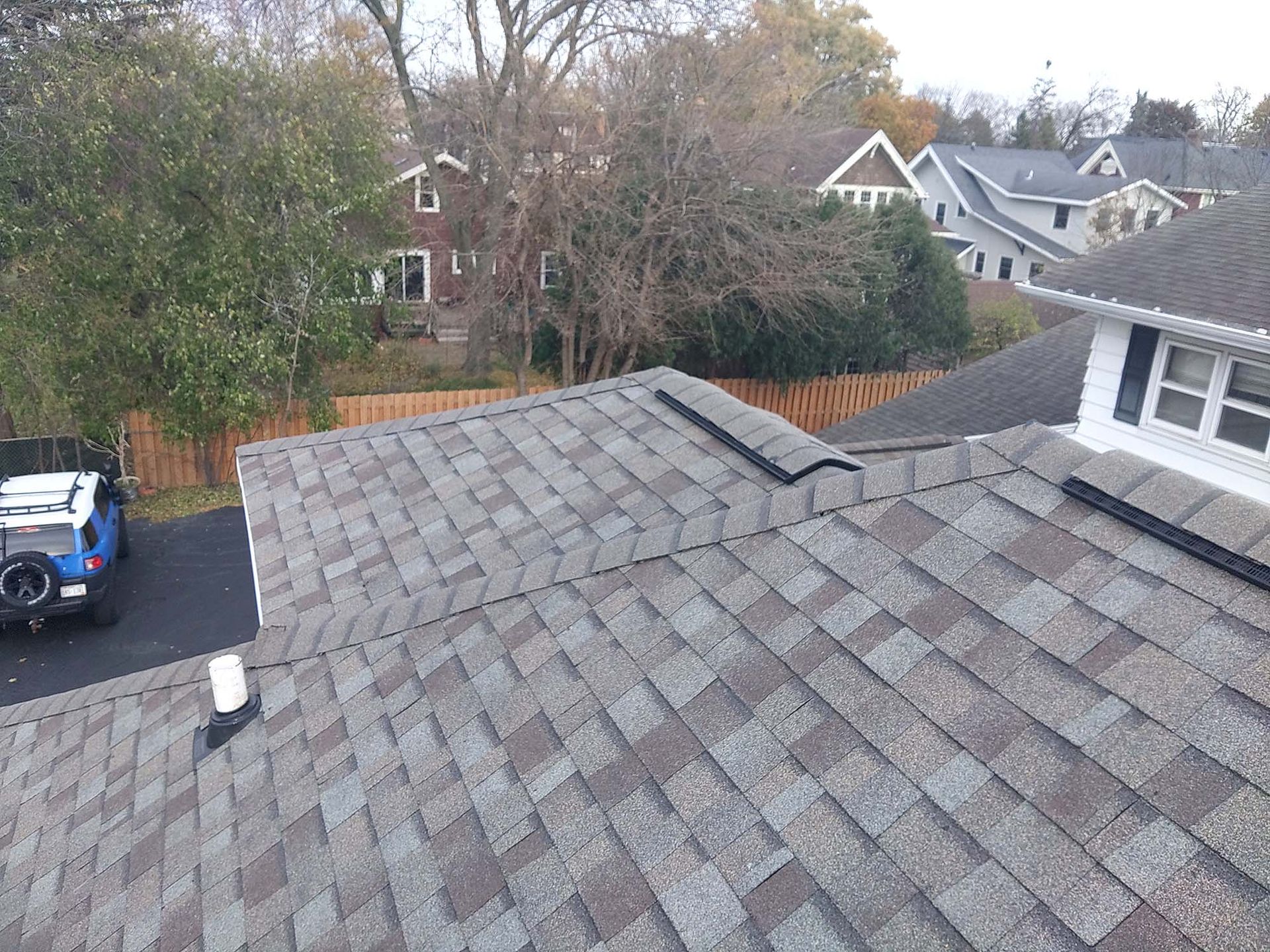 View of a shingled roof with a car and houses in the background on a sunny day.