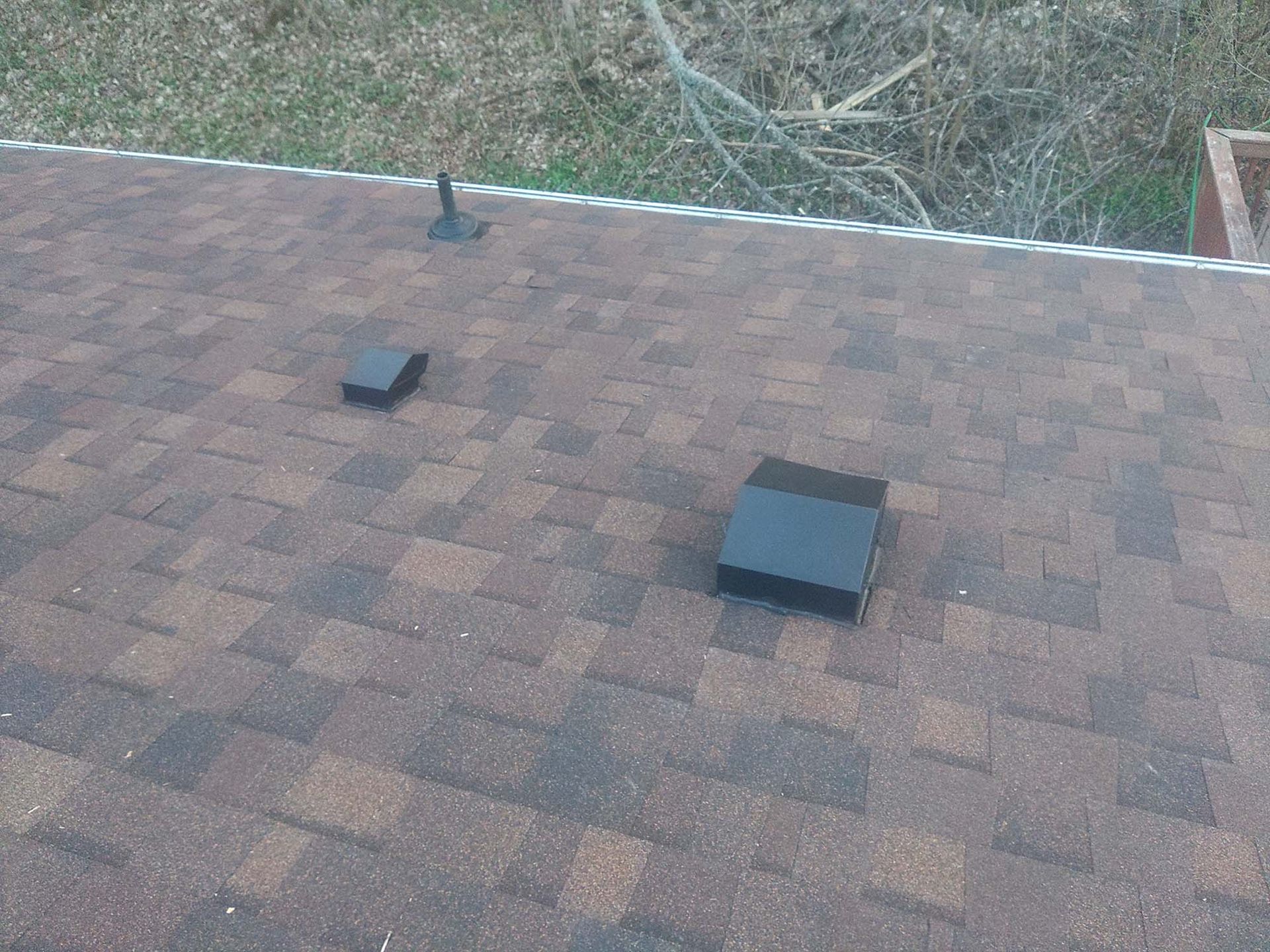 Shingled roof with two black rectangular vents and a small pipe against a grassy backdrop.
