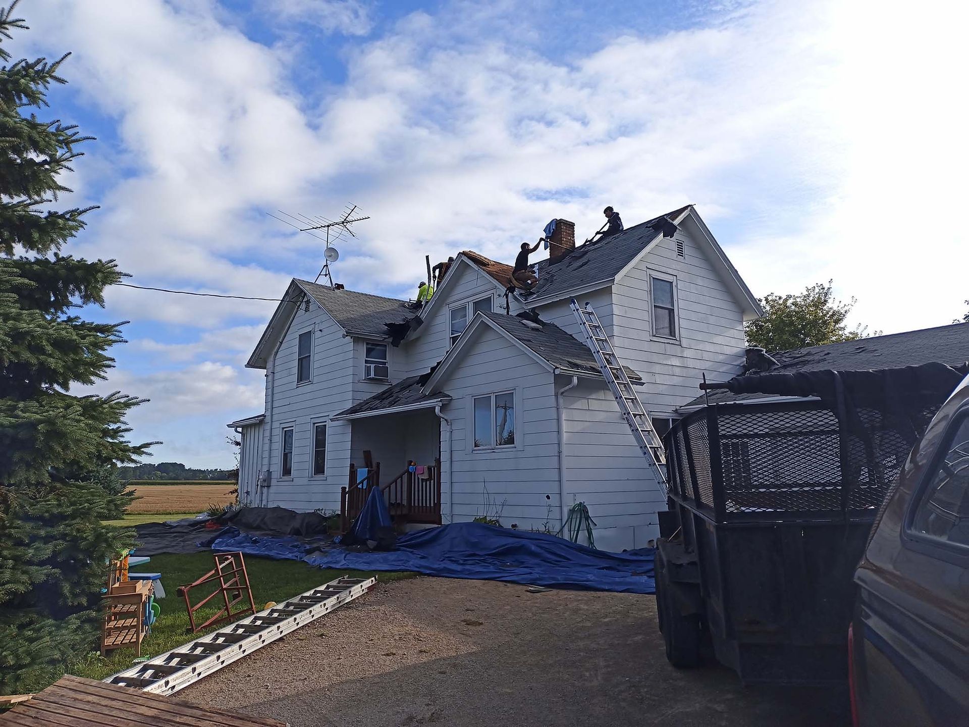 A white house with workers on the roof, blue tarp on the ground, cloudy sky.