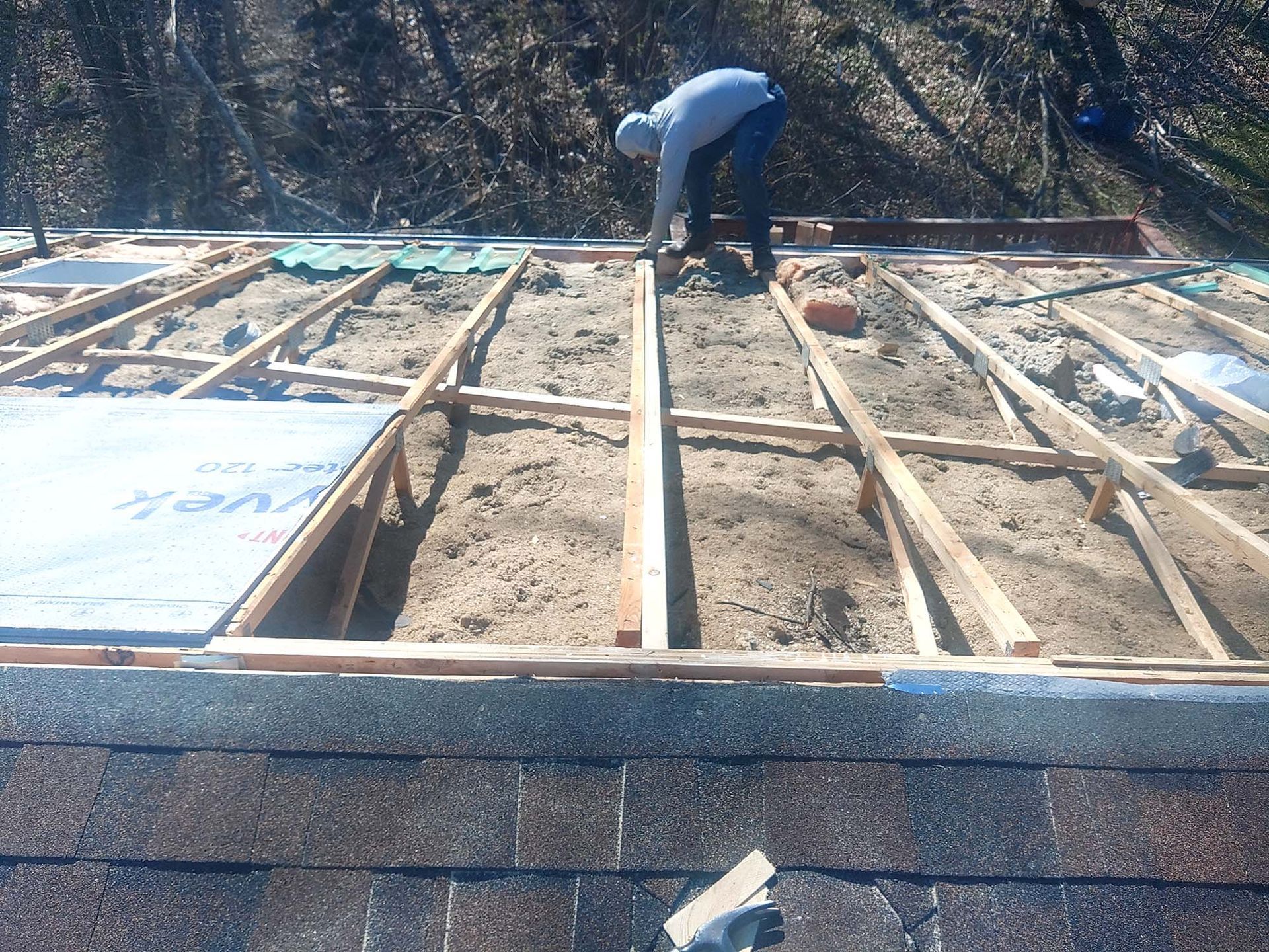 Person on roof replacing roofing materials; wood frame with insulation and existing shingles visible.