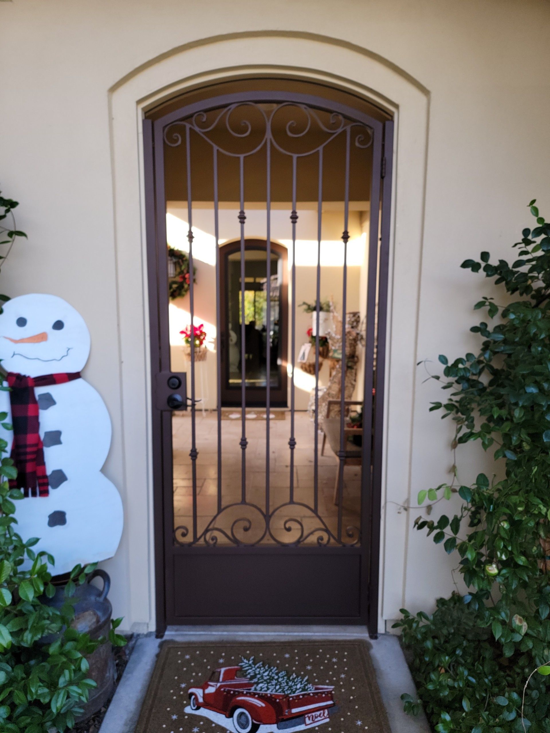 Wrought iron security door with holiday decor: snowman, welcome mat with red truck, arched opening.