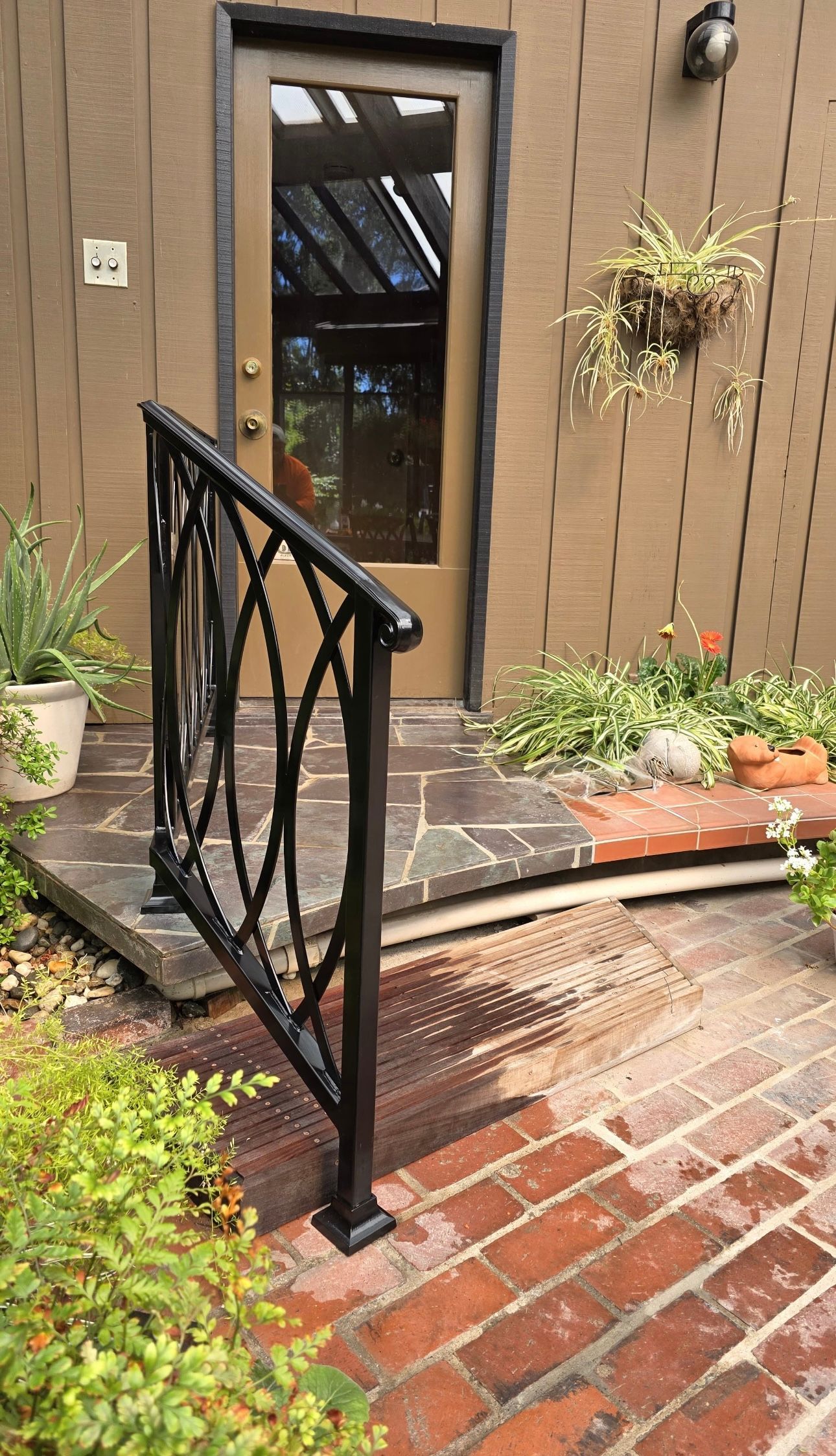 Black iron handrail beside brick steps leading to a door; potted plants nearby.