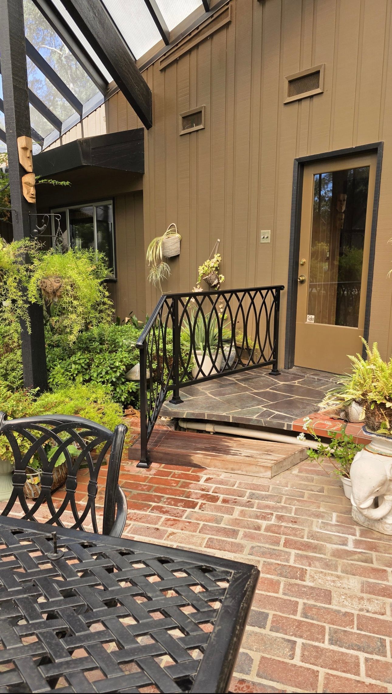Exterior view of a brick patio with wrought iron furniture, plants, and a brown building with a doorway.