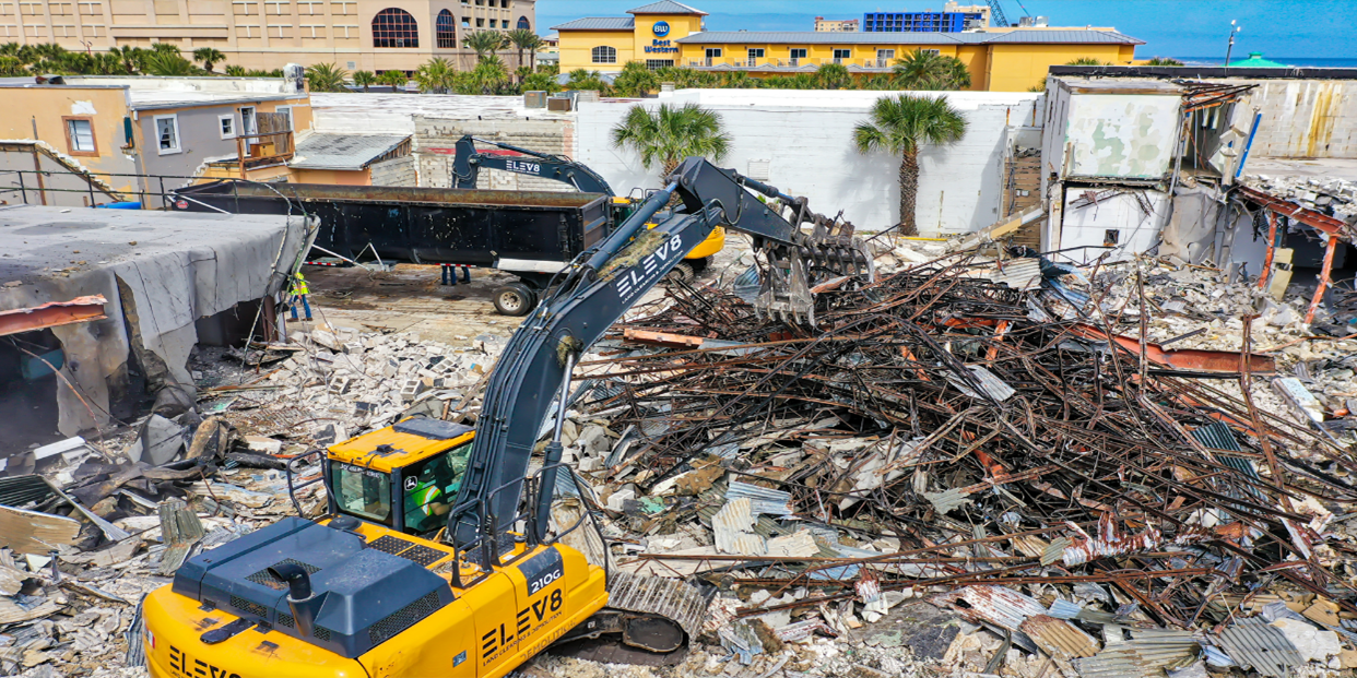 A yellow excavator demolishes a building, loading debris into a truck. Other buildings and palm trees are visible.