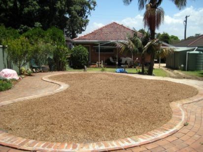 Backyard with a brick-edged gravel area, surrounded by a brick path and landscaping, with a house in the background.