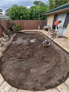 Man raking dirt within a stone-bordered area, likely a patio, next to a house with trees.