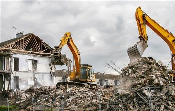 Two yellow excavators demolishing a building, creating a pile of rubble under a cloudy sky.