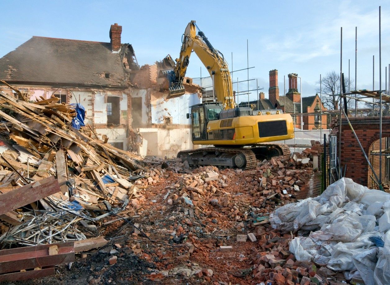 Yellow excavator demolishing a building; debris pile in foreground, other buildings in background.