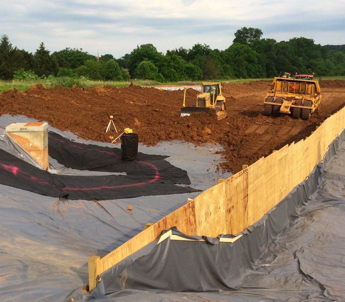 Construction site, heavy machinery, large pit lined with black material, wooden barrier.