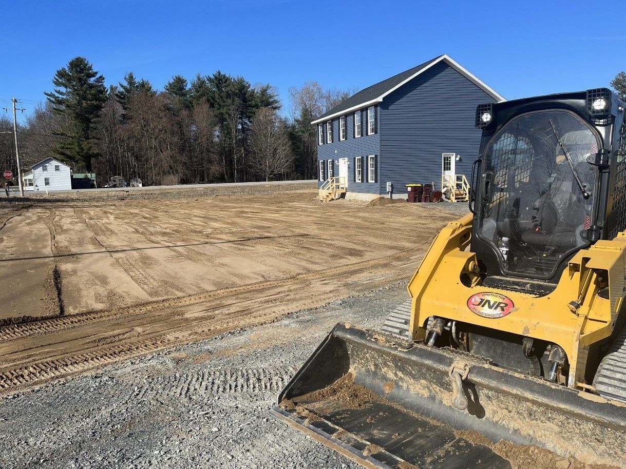 Yellow skid steer on gravel near dirt lot and blue house on a sunny day.