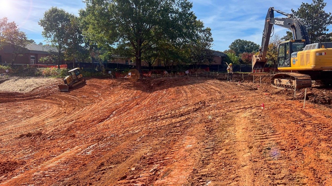 Construction site with an excavator digging into reddish-brown earth. Trees and a fence are in the background.