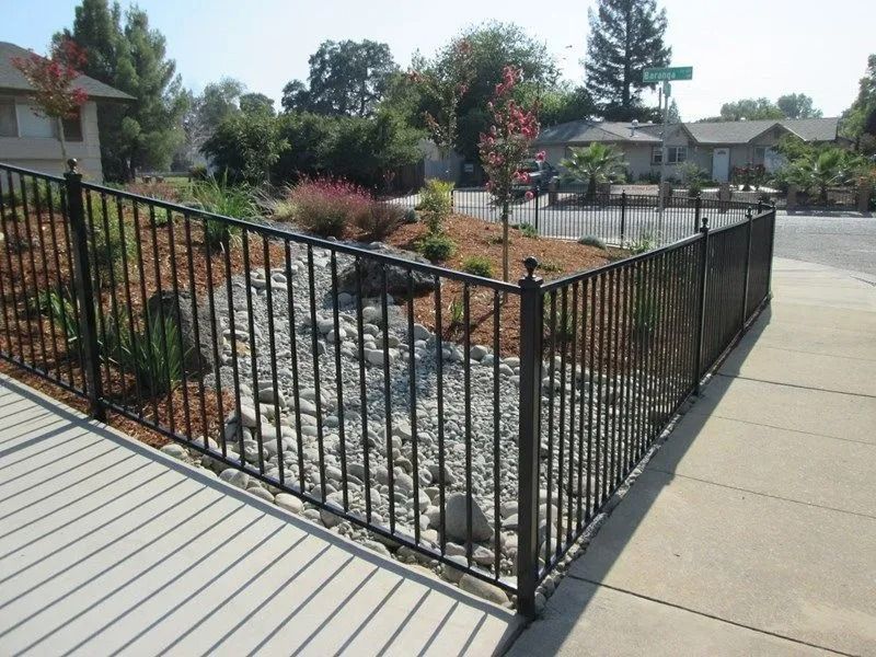 Black metal fence surrounding a landscaped rock garden along a sidewalk.