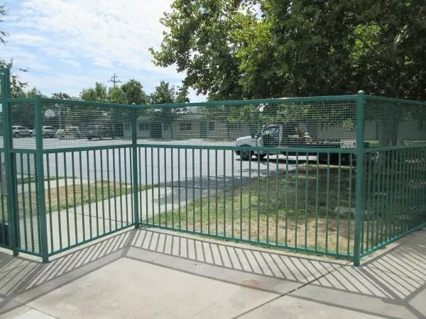Green metal security fence surrounding a parking area with trees and a building in the background.