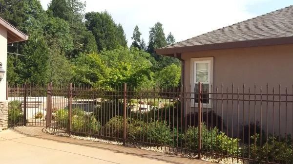 Brown wrought-iron fence surrounds a yard with greenery, next to a brown stucco house, trees in the background.