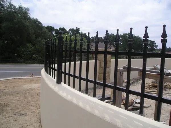 Black metal fence atop a beige curved wall. In the background is a road and trees.