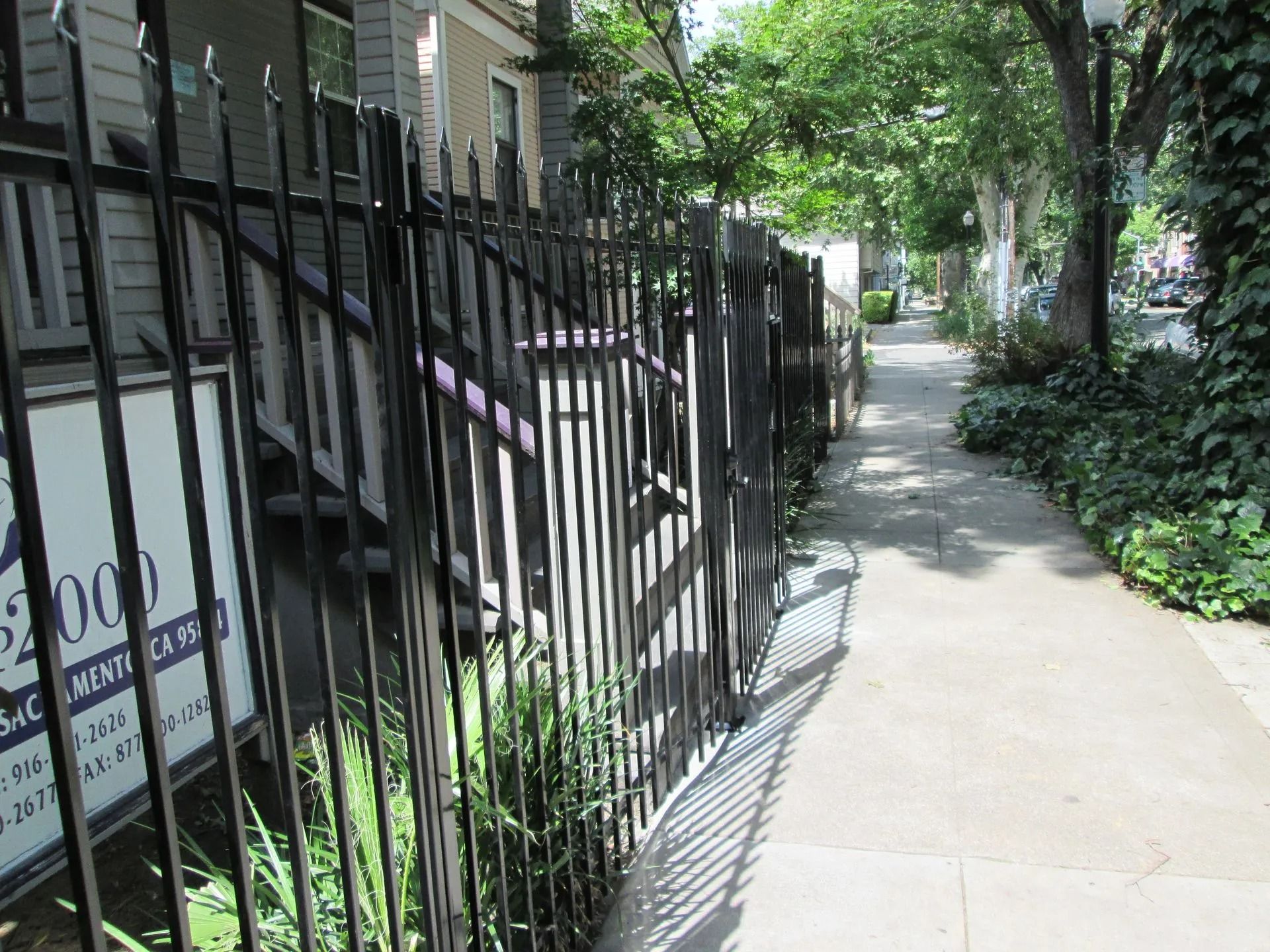 Black metal fence, sidewalk, and street with greenery and buildings.