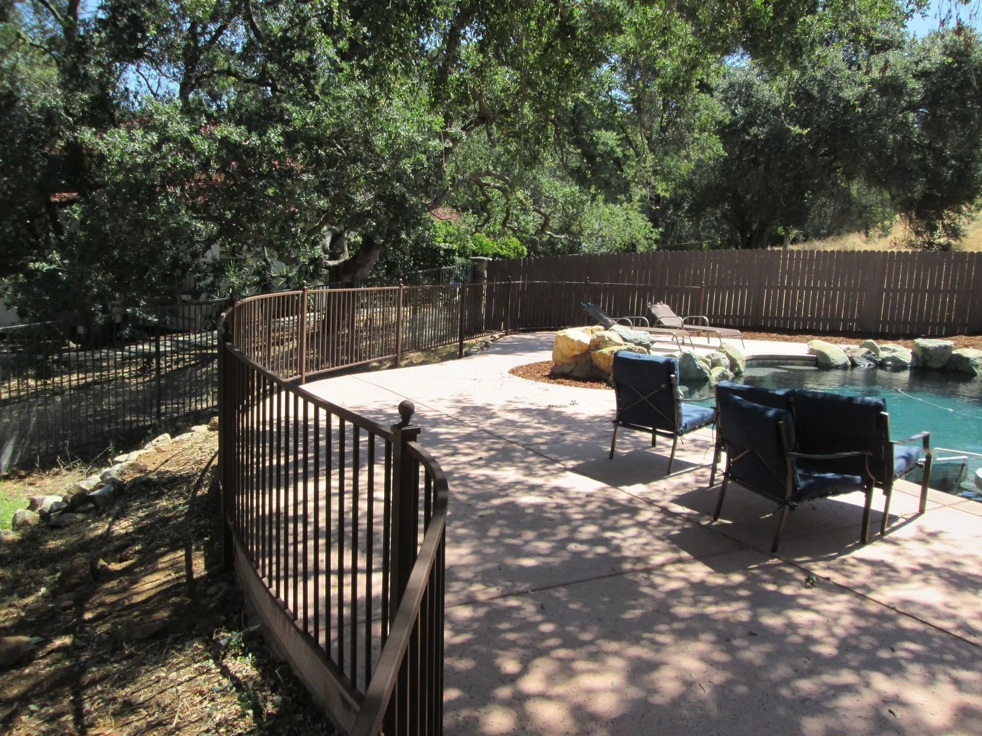 Patio with fence overlooking a pool surrounded by trees. Two chairs sit on the concrete.