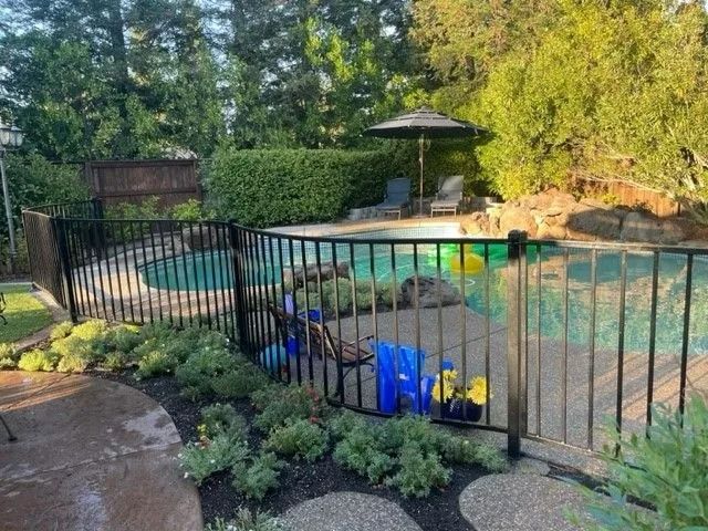 Black fence surrounds a pool with trees in the background, umbrella, lounge chair, and toys.