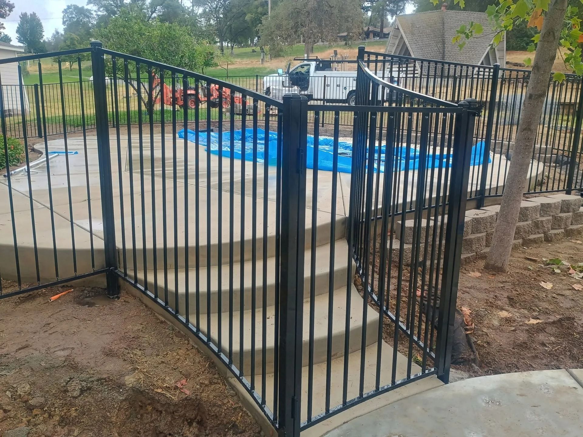 Black metal fence surrounds a swimming pool with blue cover. Stairs lead up to a gate.