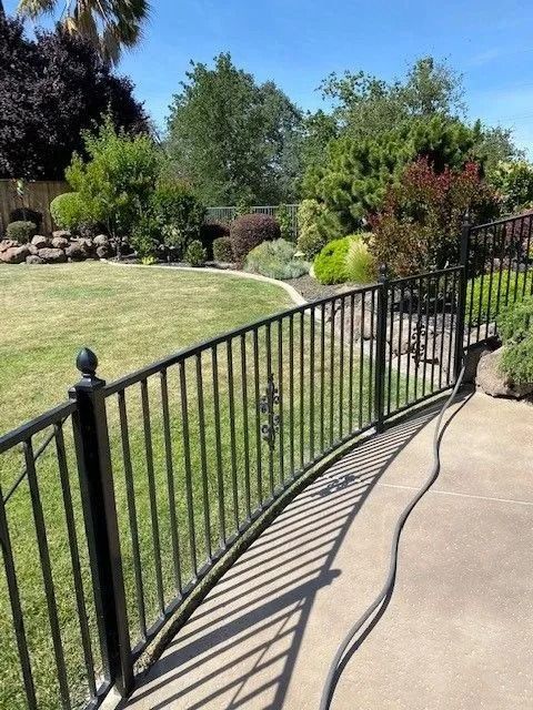 Black metal fence curves along a concrete path next to a grassy lawn and landscaped garden under a blue sky.