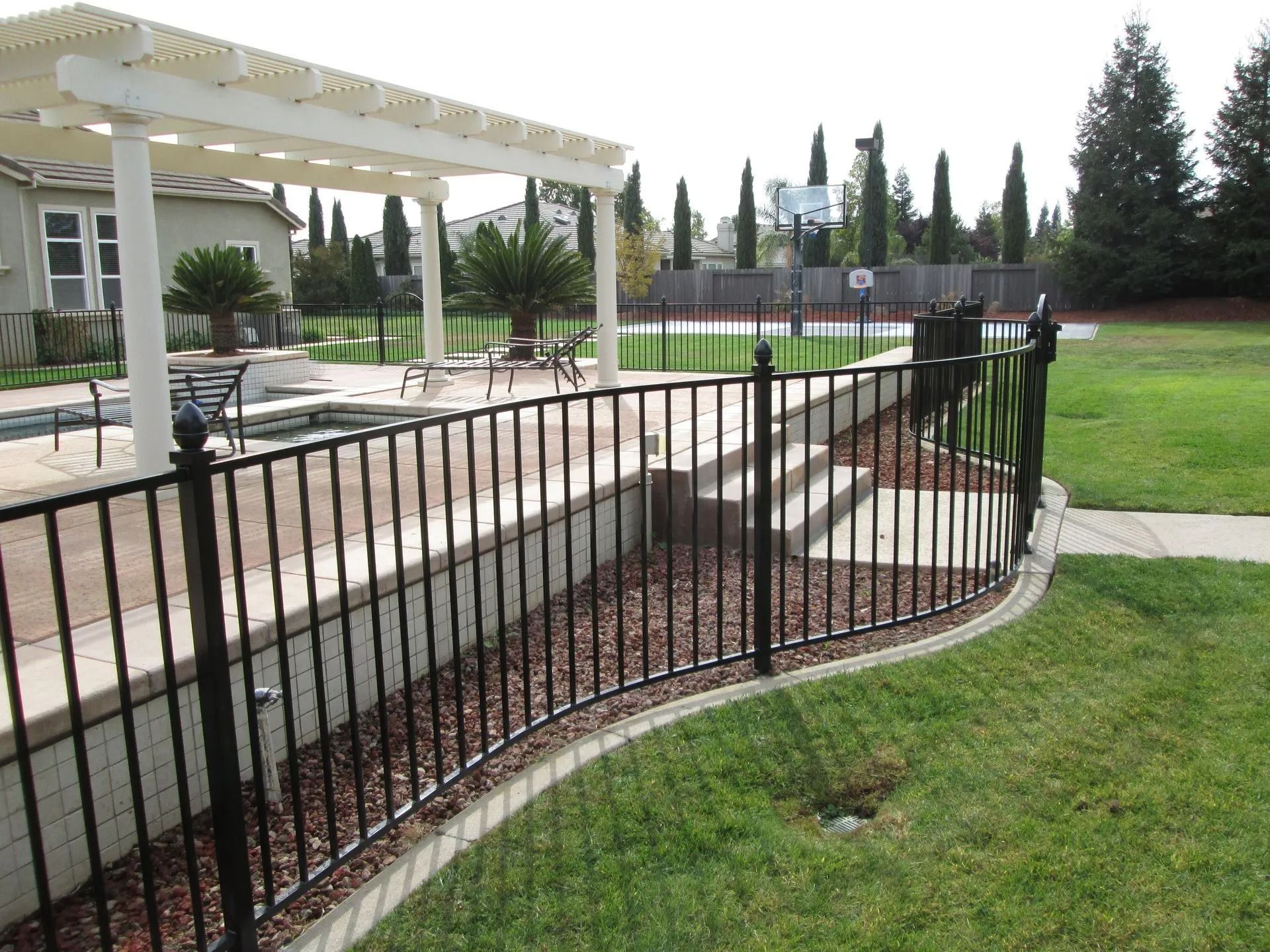 Black metal fence curving along a raised bed with a pool and pergola in the background.