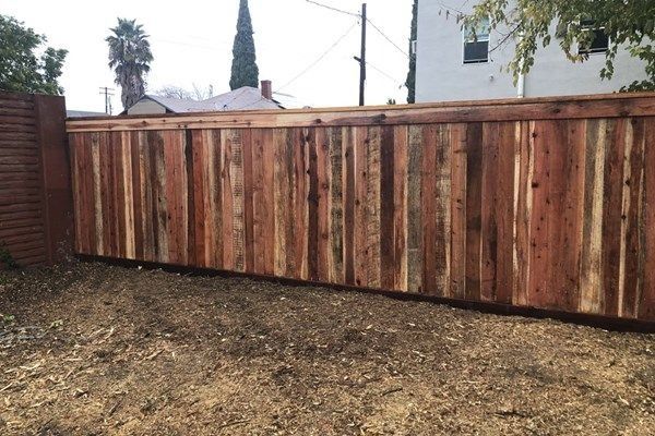 Wooden privacy fence in a yard with dirt and a few buildings visible in the background.