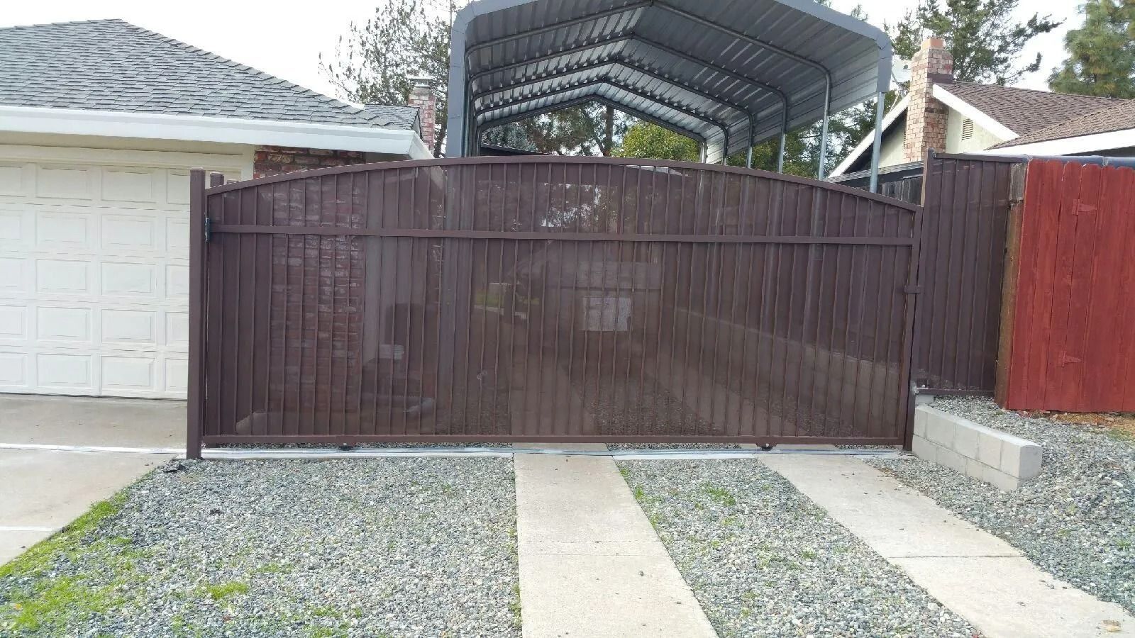 Brown metal driveway gate with a gravel driveway and a carport in the background.
