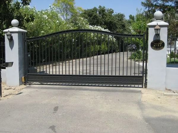 Black iron gate in front of a driveway, flanked by grey pillars with sphere tops and address plaque.