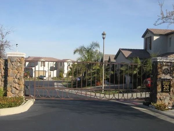 Gated community entrance with stone columns, closed metal gate, and houses in the background under a blue sky.