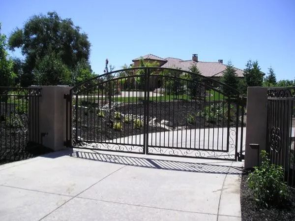 Black iron driveway gate in front of a house, set on a concrete driveway with pillars.