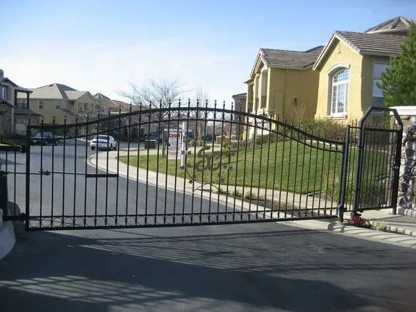 Black metal gate at the entrance to a residential community. Houses and a road are visible in the background.